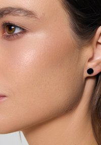 Czarna kolczyk wkrętka w lewym uchu; okrągły kształt, gładka tekstura i matowe wykończenie. Kolor skóry jasny z delikatnymi piegami i naturalnym blaskiem.