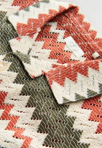 Nahaufnahme eines strukturierten, gewebten Stoffes mit Zickzackmustern in Grün, Weiß und Orange auf einem Kleidungsstück mit Kragen und Knopf.