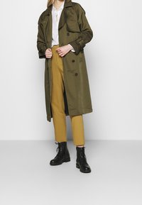 Trench coat verde oliva con bottoni doppi, indossato sopra una camicia bianca e pantaloni gialli. Stivali da combattimento neri con lacci completano l'outfit.