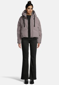 Frau in einer mauvefarbenen gepolsterten Kapuzenjacke, schwarzem enganliegenden Top, schwarzen schmalen Hosen und schwarzen Stiefeletten, stehend vor einem schlichten weißen Hintergrund.