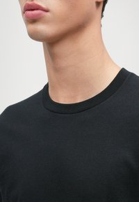 Μαύρο βαμβακερό μπλουζάκι με λαιμόκοψη και κοντά μανίκια, που διαθέτει λεία υφή και ραφή στο λαιμό.