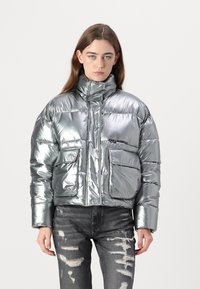Sølv pufferjakke med høy krage, som har to frontlommer og en cropped design. Glatt, reflekterende stoff med sydde segmenter.