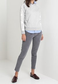 Vrouw draagt een grijze GANT hoodie met logostrepen op de mouwen, een lichtblauw overhemd, grijze broek en donkerbruine loafers, staand tegen een effen achtergrond.