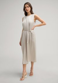 Lichtbeige, mouwloze midi-jurk met een strikceintuur in de taille, met zachte plooien en een soepele, vloeiende textuur. Heldere sandalen met blokhak.