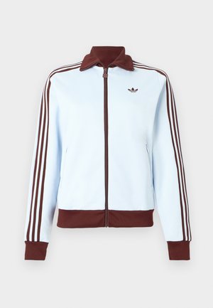 Lichtblauwe jas met ritssluiting, bruine kraag, manchetten en zoom, en drie bruine strepen langs elke mouw, met een klein bruin logo op de borst.