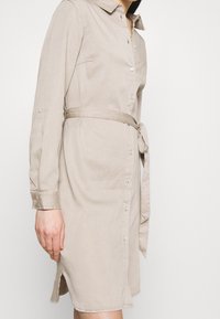 Beige napitettava mekko, joka on valmistettu pehmeästä kankaasta. Mekossa on kaulus, pitkät hihat, vyötäröllä matching-vyö ja sivuaukot.