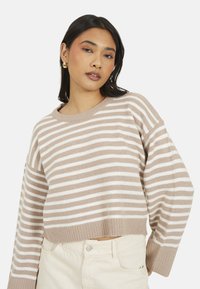 Giovane donna con capelli scuri, che indossa orecchini a cerchio dorati, un maglione a righe beige e bianche e pantaloni chiari, in posa contro uno sfondo neutro.