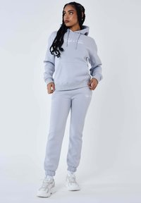 Lichtgrijze hoodie en bijpassende sweatpants van zachte stof. De hoodie heeft een voorzak en een trekkoord; de broek heeft elastische boorden en logo-detail.