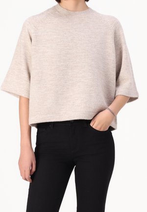 Personne portant un pull en maille beige à manches courtes et un pantalon noir, avec une main dans la poche et l'autre détendue le long du corps.
