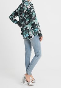 Donna che indossa una blusa nera a fiori con maniche lunghe, jeans skinny azzurri chiari e scarpe con tacco alto argentate, in piedi su uno sfondo semplice.
