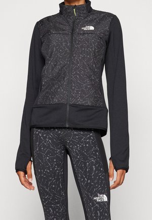 Personne portant une veste noire The North Face et des leggings avec un motif abstrait éclaboussé gris, debout devant un fond clair.