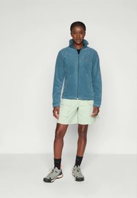 Veste en polar bleu avec fermeture éclair devant et col haut, associée à un short cargo vert clair et des baskets grises. Design simple avec des éléments pratiques.