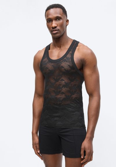 Άνδρας με μαύρο ματιωτό αμάνικο μπλουζάκι με γεωμετρικά σχέδια και μαύρο σορτς, στέκεται μπροστά σε ένα απλό ανοιχτό φόντο.