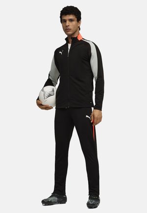 Chándal deportivo negro con acentos grises, que incluye una chaqueta con cremallera y pantalones ajustados. El modelo sostiene un balón de fútbol blanco y lleva tacos.