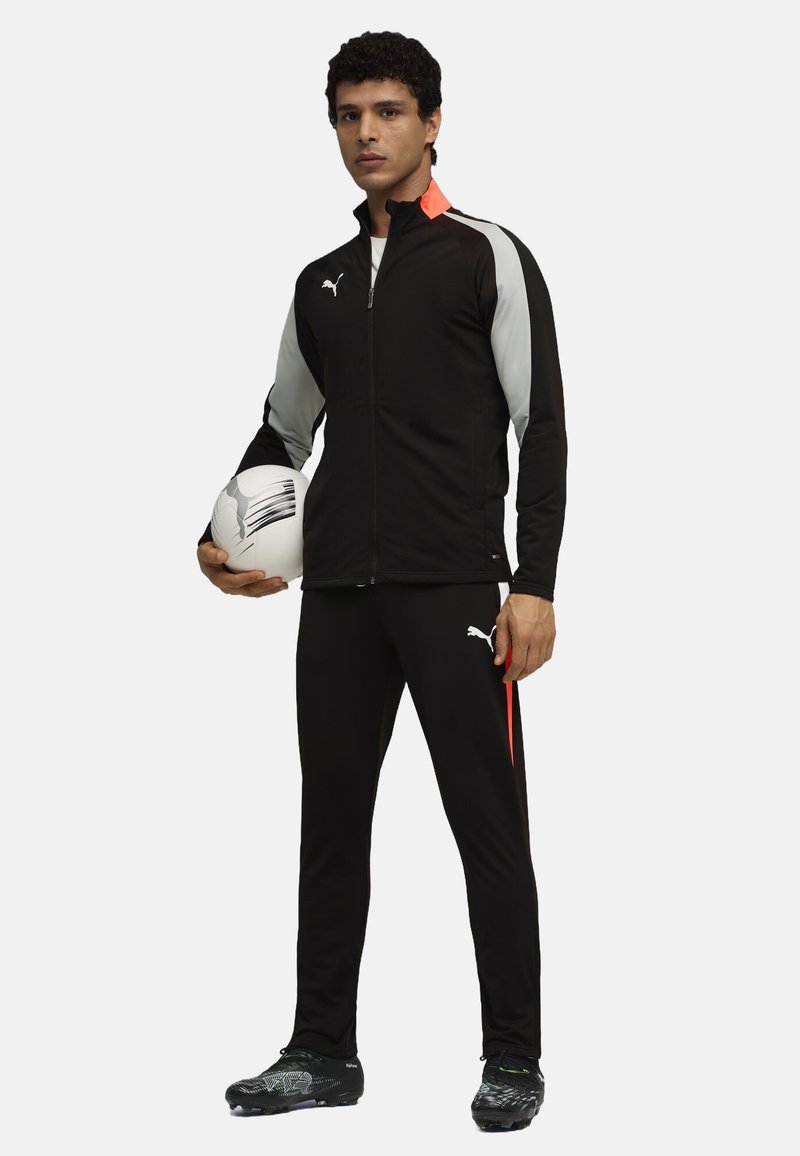 Tuta sportiva nera con accenti grigi, caratterizzata da un giubbotto con zip e pantaloni aderenti. Il modello tiene in mano un pallone da calcio bianco, indossando delle scarpe da calcio.