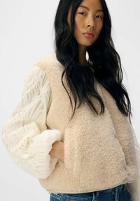 Beige fleece vest met een zachte textuur, gelaagd over een witte, gerimpelde, doorzichtige blouse met lange mouwen. Denim jeans maken de look compleet.