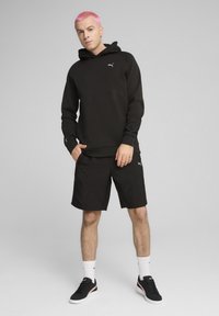 Sort hættejakke med kængurulomme, parret med sorte shorts. Sorte sneakers med hvide såler. Modellen har pink hår og står op.