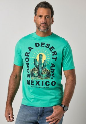 Mężczyzna w turkusowej koszulce z dużym nadrukiem kaktusa, słońca i krajobrazu pustyni oraz napisem „Sonora Desert Adv. Mexico.”