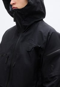 Μαύρο αδιάβροχο μπουφάν με ψηλό γιακά, ρυθμιζόμενη κουκούλα και πολλαπλές τσέπες με φερμουάρ. Διαθέτει λεία υφή και κομψό σχεδιασμό.