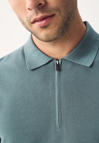 Poloshirt mit Reißverschluss in Schieferblau, mit einer Strickstruktur und einem umklappbaren Kragen mit kleinem Reißverschluss am Hals.