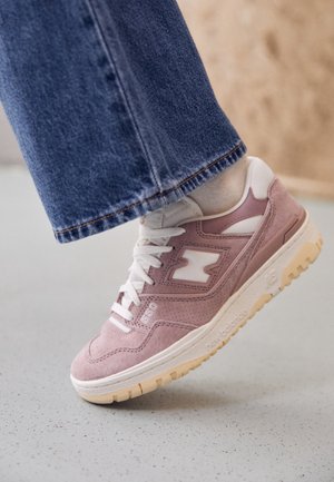 Light pink suede sneaker with white accents, rounded toe, textured upper, and beige rubber sole. Features laces and logo on the side.