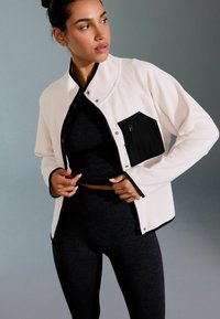 Weißes Fleecejäckchen mit schwarzen Akzenten und einer Reißverschlusstasche, getragen über einem schwarzen Oberteil und Leggings, das ein modernes, figurnahes Design zeigt.