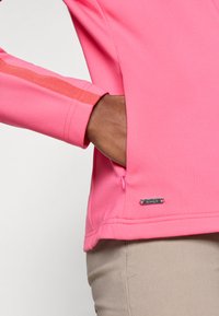 Fitted pink sweatshirt with long sleeves, textured fabric, side zipper pocket, and a metallic logo accent near the hem.
