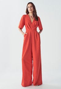 Rode jumpsuit met een diepe V-hals, korte pofmouwen en wijde pijpen. Bevat zijzakken en heeft een gestructureerde stof.