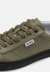 Ελιές πράσινα δερμάτινα αθλητικά παπούτσια με καφέ υφασμάτινες κορδόνες, μαύρη καουτσούκ σόλα και λευκή ετικέτα λογότυπου. Διαθέτει υφή επιφάνειας και λεία στοιχεία.