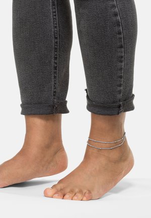 Knöchel mit zwei dünnen silbernen Kettenanklets, neben hochgekrempelten dunkelgrauen Jeans, die den baren Fuß und die Zehen auf einem neutralen Hintergrund zeigen.
