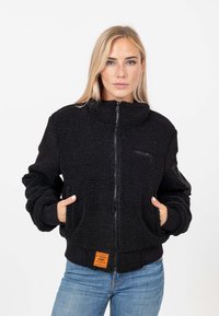 Giubbotto bomber in pile nero con colletto alto, chiusura con zip, tasche laterali e polsini a coste. Presenta un'etichetta arancione del marchio sull'orlo.