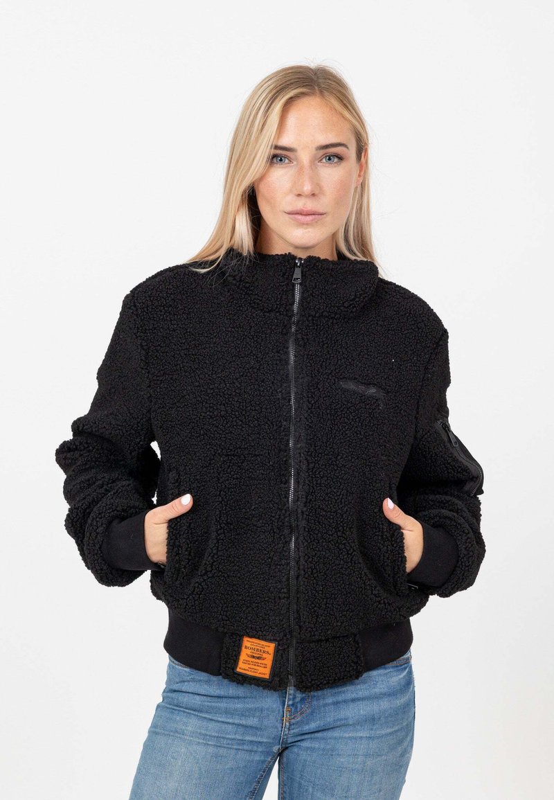 Giubbotto bomber in pile nero con colletto alto, chiusura con zip, tasche laterali e polsini a coste. Presenta un'etichetta arancione del marchio sull'orlo.