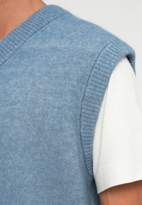 Pulôver de malha sem mangas azul claro usado sobre uma camisa de manga curta branca na parte superior do torso de uma pessoa.