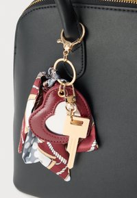 Μαύρη τσάντα χειρός με ομαλή υφή, διακοσμημένη με χρυσό κούμπωμα, μπρελόκ καρδιά σε μπορντό χρώμα και φουλάρι με σχέδια.