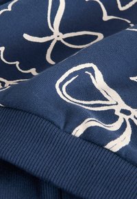 Blått tyg med vita blommönster och rosettmönster, med en strukturerad yta och ribbade ärmslut. Designen är tydligt framträdande.