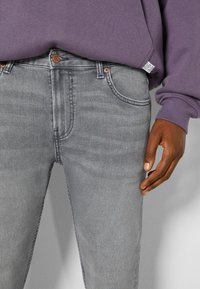 Primer plano de una persona que lleva unos vaqueros de mezclilla grises y una sudadera morada con una etiqueta visible en la costura lateral.