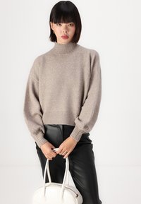 Beige, rövidített pulóver magas bordás nyakkal és ballon ujjakkal, fekete bőrnadrággal és fehér táskával párosítva. Puha textúra jellemző.