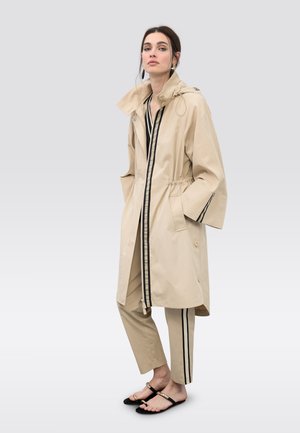 Impermeable beige con cremallera frontal, cintura elástica y rayas negras. Combinado con pantalones beige con rayas laterales y sandalias negras.