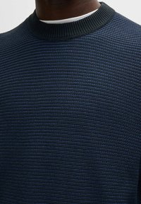 Gros plan d'une personne portant un pull en maille texturée bleu foncé sur un sous-vêtement blanc, montrant le col côtelé et le motif en maille.