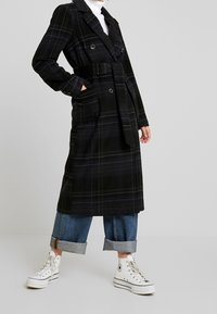 Persona che indossa un lungo cappotto a scacchi scuro con cintura, jeans azzurri arrotolati, sneakers bianche alte e un dolcevita bianco su uno sfondo semplice.