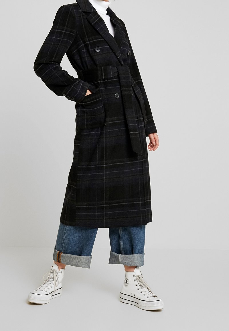 Persona che indossa un lungo cappotto a scacchi scuro con cintura, jeans azzurri arrotolati, sneakers bianche alte e un dolcevita bianco su uno sfondo semplice.