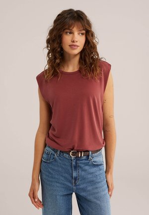 Kobieta o kręconych brązowych włosach, w bezrękawnym bordowym topie wpuszczonym w niebieskie jeansy z czarnym paskiem na jednolitym tle.