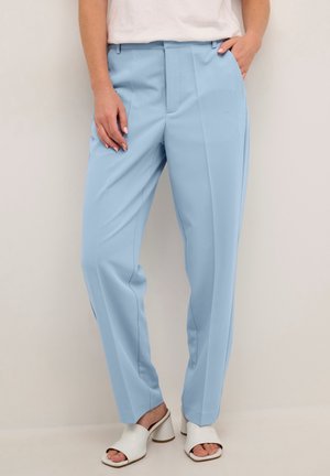 Femme portant un pantalon bleu clair cintré et des sandales à talons blocs blanches à bout ouvert, debout avec une main dans la poche.