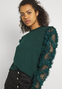 Dark green long-sleeve top with sheer mesh sleeves adorned with layered teal ruffles. Fitted silhouette, paired with a black button-up skirt.