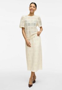 Vestido midi de un beige claro con mangas cortas abullonadas, que presenta un patrón de corte en cuadrícula. Complementado con tacones de punta en un color marrón intenso.
