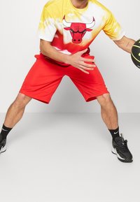 Czerwone krótkie spodenki sportowe w połączeniu z koszulką tie-dye w żółto-białe paski z czarno-czerwonym nadrukiem byka, noszone z czarnymi trampkami i skarpetkami.