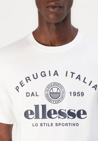 Bílý bavlněný tričko s grafickými prvky v námořnické modři, včetně kulatého vzoru a textu "Perugia Italia" a "ellesse" napsaného tučným písmem. Viditelný kulatý výstřih.