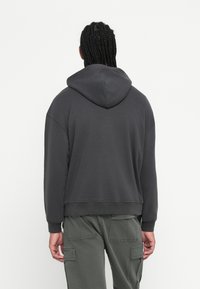 Άτομο με πλεξούδες που φοράει σκούρο γκρι hoodie και ελαιόχρωμο παντελόνι cargo, εμφανίζεται από πίσω ενάντια σε λευκό φόντο.