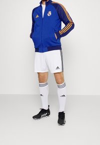 Kungligt blå zip-up jacka med orange detaljer, ihop med vita shorts och svarta fotbollsskor. Adidas-logotyp på klädernas och skornas material.