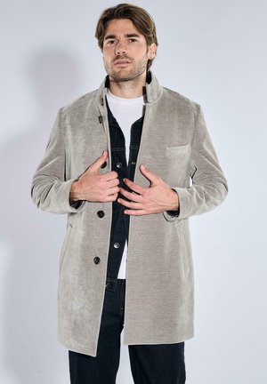 Man wearing a light grey buttoned coat over a black jacket and white shirt, standing against a plain light background.